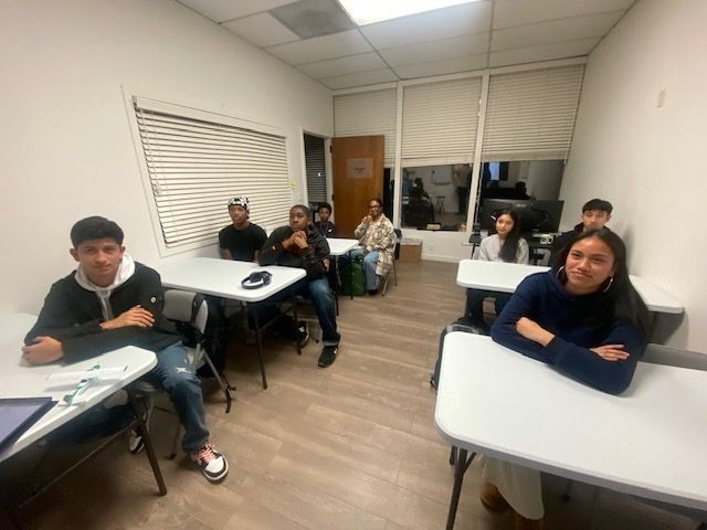 Small classroom with students seated at desks, some using laptops and others listening attentively.