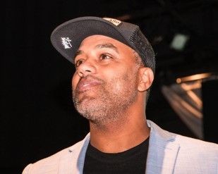 Man wearing a cap and light-colored blazer looking upwards against a dark background.