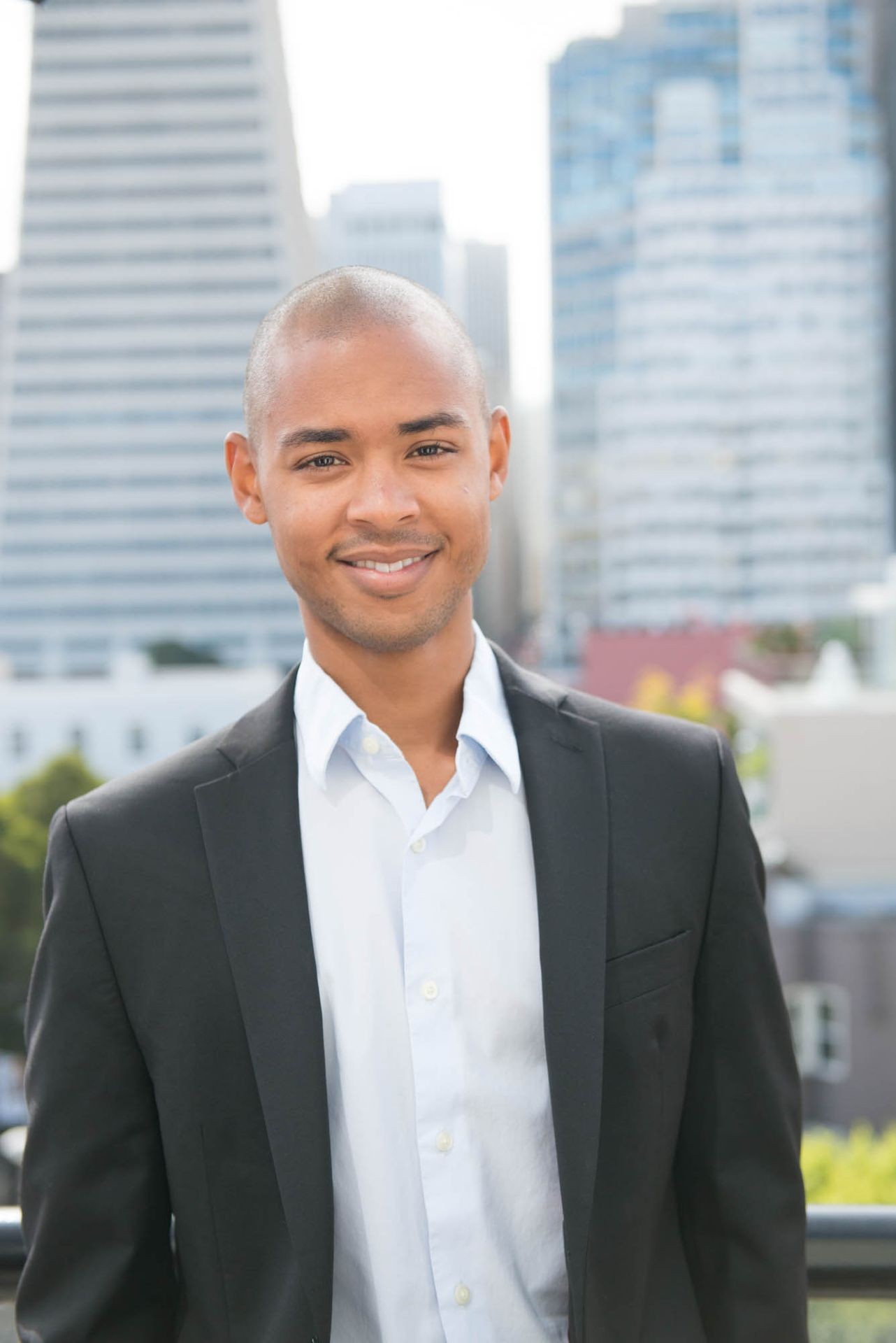 Person in a black suit jacket and white shirt standing outdoors with modern buildings in the background.