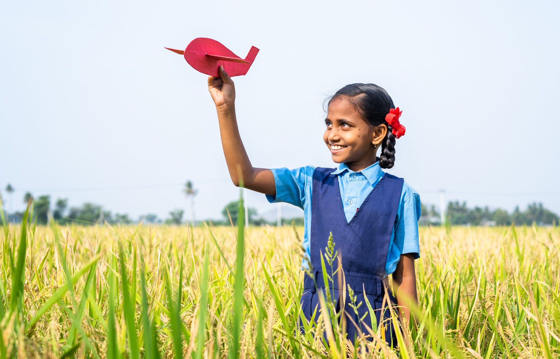 Happy village girl kid with school uniform playing using toy cardboard aeroplane at paddy field - concept of childhood dream, aspirations and freedom.